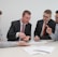 Four men in business attire are engaged in a discussion around a table. One man appears to be explaining something while pointing at some documents in front of them. The others are listening attentively, and one holds a pen and a mobile device. The setting is a plain office environment.