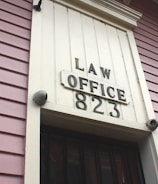 A close-up view of an exterior building wall with horizontal paneling painted in a soft pink color. The central focus is a large rectangular plaque stating 'LAW OFFICE 823' in bold letters. The paneling and design suggest a historical or vintage style.