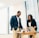 Two people are standing in an office space near a table with a laptop and papers. They are dressed in business attire and appear to be having a discussion.