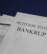 Documents are laid out on a surface, with the largest sheet displaying the text 'Petition to File for Bankruptcy'. Several forms and paperwork are visible underneath, suggesting a formal or legal context.