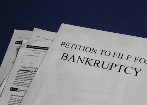 Documents are laid out on a surface, with the largest sheet displaying the text 'Petition to File for Bankruptcy'. Several forms and paperwork are visible underneath, suggesting a formal or legal context.