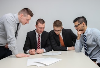 A group of four men in business attire are gathered around a table, examining documents laid out in front of them. They appear to be engaged in a discussion or meeting, with focused expressions and attentive body language.