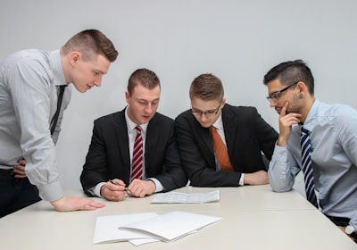 A group of four men in business attire are gathered around a table, examining documents laid out in front of them. They appear to be engaged in a discussion or meeting, with focused expressions and attentive body language.