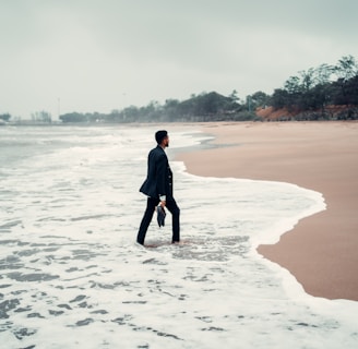 A person in business attire stands with one foot in the ocean surf on a deserted beach, holding a briefcase. The sky is overcast, and the surrounding scenery is lined with distant trees and greenery.