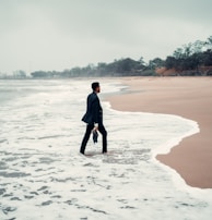 A person in business attire stands with one foot in the ocean surf on a deserted beach, holding a briefcase. The sky is overcast, and the surrounding scenery is lined with distant trees and greenery.