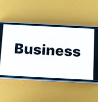 A smartphone lying on a yellow surface displays the word 'Business' in bold black text on its screen.