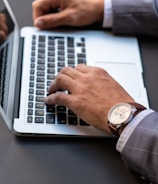 A person wearing a business suit types on a laptop keyboard. They have a wristwatch with a brown leather strap on their left wrist. The scene captures a professional setting, possibly in an office.