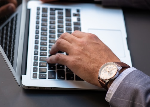 A person wearing a business suit types on a laptop keyboard. They have a wristwatch with a brown leather strap on their left wrist. The scene captures a professional setting, possibly in an office.