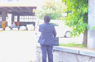 A person wearing a dark suit is standing on a sidewalk facing away and holding a briefcase. There is a stone ledge beside them, and the background includes greenery on the right and a lightly trafficked urban area with a white vehicle in the distance.