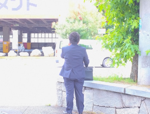 A person wearing a dark suit is standing on a sidewalk facing away and holding a briefcase. There is a stone ledge beside them, and the background includes greenery on the right and a lightly trafficked urban area with a white vehicle in the distance.