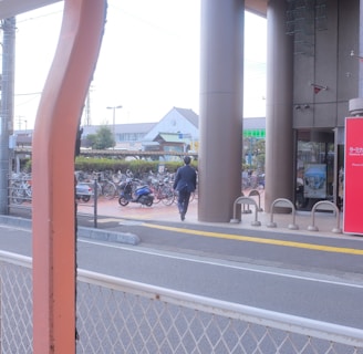 A person in business attire is walking toward a building with tall pillars. There are bicycles parked in a bike rack area, and a red sign with text near the entrance. A road with a white metal fence is in the foreground, and buildings are visible in the background.