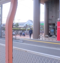 A person in business attire is walking toward a building with tall pillars. There are bicycles parked in a bike rack area, and a red sign with text near the entrance. A road with a white metal fence is in the foreground, and buildings are visible in the background.