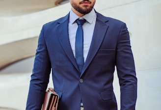 A man in a business suit stands confidently holding a leather briefcase. The background features a modern architectural wall with curved lines. The man has a neatly groomed beard and styled hair, wearing a dark blue suit with a matching tie and a striped shirt.