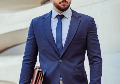 A man in a business suit stands confidently holding a leather briefcase. The background features a modern architectural wall with curved lines. The man has a neatly groomed beard and styled hair, wearing a dark blue suit with a matching tie and a striped shirt.