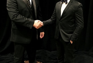 Two men dressed in formal black suits are shaking hands in an indoor setting with a dark curtain backdrop. The man on the left is holding a piece of paper, and both have a professional demeanor.