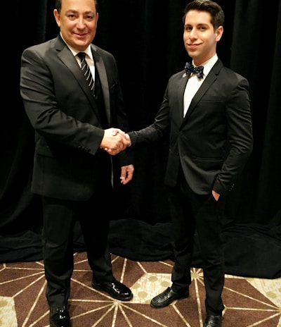 Two men dressed in formal black suits are shaking hands in an indoor setting with a dark curtain backdrop. The man on the left is holding a piece of paper, and both have a professional demeanor.