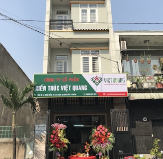 A multi-story building with a business on the ground floor displaying a sign for an architectural company. The facade includes balconies with railings. In front of the entrance, there is a table covered with a red cloth, adorned with decorative floral arrangements. Motorbikes are parked along the sidewalk and there are some potted plants on a ledge above the entrance.