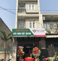 A multi-story building with a business on the ground floor displaying a sign for an architectural company. The facade includes balconies with railings. In front of the entrance, there is a table covered with a red cloth, adorned with decorative floral arrangements. Motorbikes are parked along the sidewalk and there are some potted plants on a ledge above the entrance.