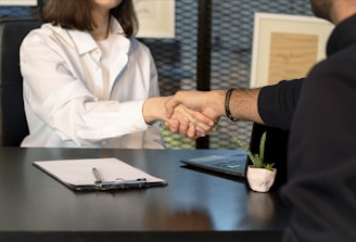 Two people sitting at a desk are shaking hands over paperwork, likely indicating the conclusion of a business deal or an agreement. One person is wearing a white shirt and the other is dressed in dark clothing. Other items present include a laptop, a small potted plant, and a clipboard with a pen.