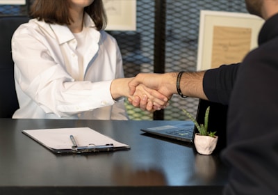 Two people sitting at a desk are shaking hands over paperwork, likely indicating the conclusion of a business deal or an agreement. One person is wearing a white shirt and the other is dressed in dark clothing. Other items present include a laptop, a small potted plant, and a clipboard with a pen.