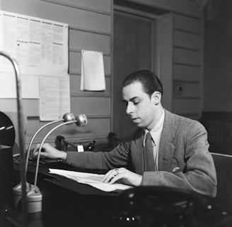 A man wearing a suit sits at a desk, concentrating on the documents in front of him. The desk has a microphone, a telephone, and some papers. The background includes a wall with several sheets of paper posted on it and a door.