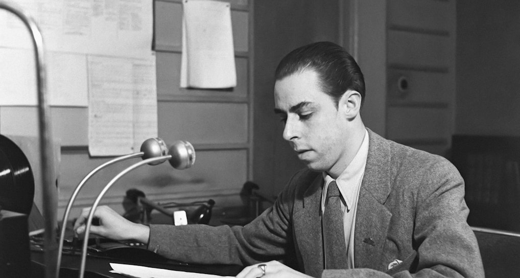 A man wearing a suit sits at a desk, concentrating on the documents in front of him. The desk has a microphone, a telephone, and some papers. The background includes a wall with several sheets of paper posted on it and a door.