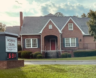 A traditional red brick building with a gabled roof and white-framed windows. The front entrance features an arched doorway and is flanked by well-maintained shrubs. A sign in the foreground reads 'Morris & Dean, Trial Lawyers & Counselors,' displaying the phone number and a digital clock showing 6:31.