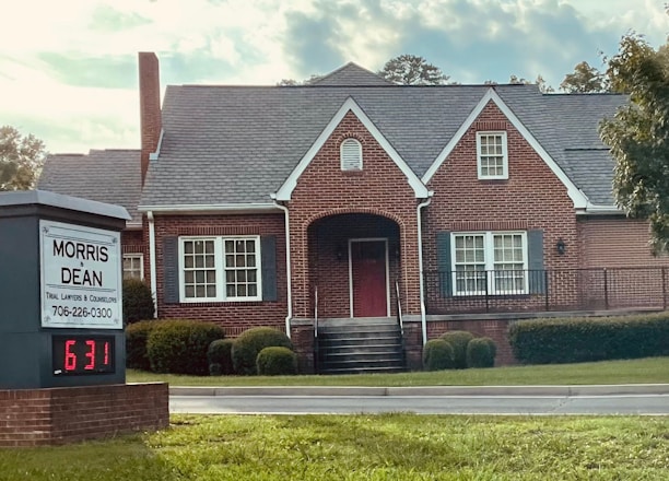 A traditional red brick building with a gabled roof and white-framed windows. The front entrance features an arched doorway and is flanked by well-maintained shrubs. A sign in the foreground reads 'Morris & Dean, Trial Lawyers & Counselors,' displaying the phone number and a digital clock showing 6:31.