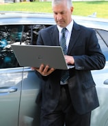 A man in a business suit is standing next to a car while using a laptop. The background includes a grassy area and a building with a garage door. The man appears focused on the task at hand.