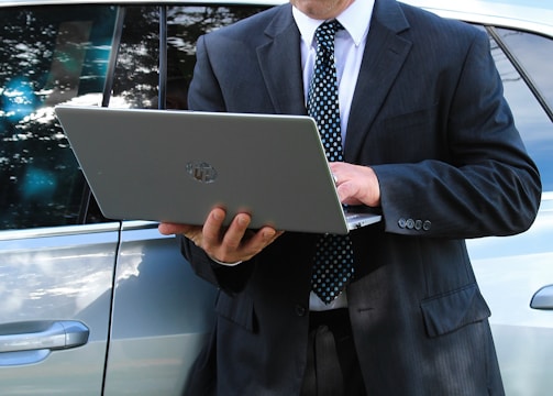 A man in a business suit is standing next to a car while using a laptop. The background includes a grassy area and a building with a garage door. The man appears focused on the task at hand.