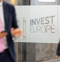 Two individuals in business attire are standing near glass doors with the phrase 'Invest Europe' etched on them. One person is holding a smartphone.