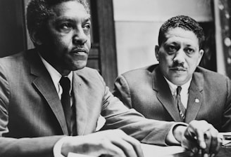 Two men in suits sit at a table, appearing engaged and focused on an ongoing discussion or meeting. Papers and documents are present on the table, indicating a formal setting.