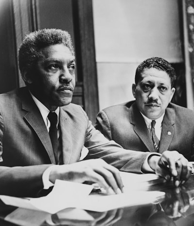 Two men in suits sit at a table, appearing engaged and focused on an ongoing discussion or meeting. Papers and documents are present on the table, indicating a formal setting.