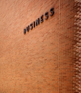 A large brick wall with the word 'BUSINESS' prominently displayed in black letters. The wall is lit by natural daylight, casting gentle shadows along its surface. The ground at the base of the wall has some patches of grass and is bordered by a concrete sidewalk. There are three metal fixtures attached to the lower part of the wall.