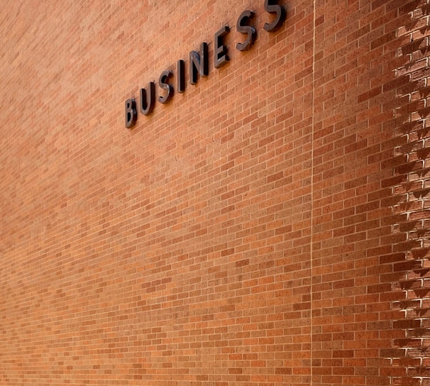 A large brick wall with the word 'BUSINESS' prominently displayed in black letters. The wall is lit by natural daylight, casting gentle shadows along its surface. The ground at the base of the wall has some patches of grass and is bordered by a concrete sidewalk. There are three metal fixtures attached to the lower part of the wall.